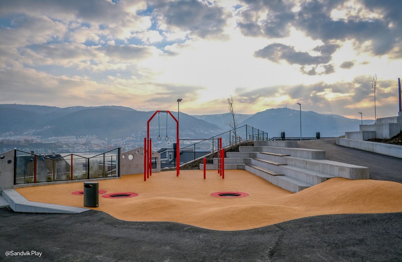 Sandfarbene Fläche mit roten Trainingsgeräten vor einer bergigen Küstenlandschaft.