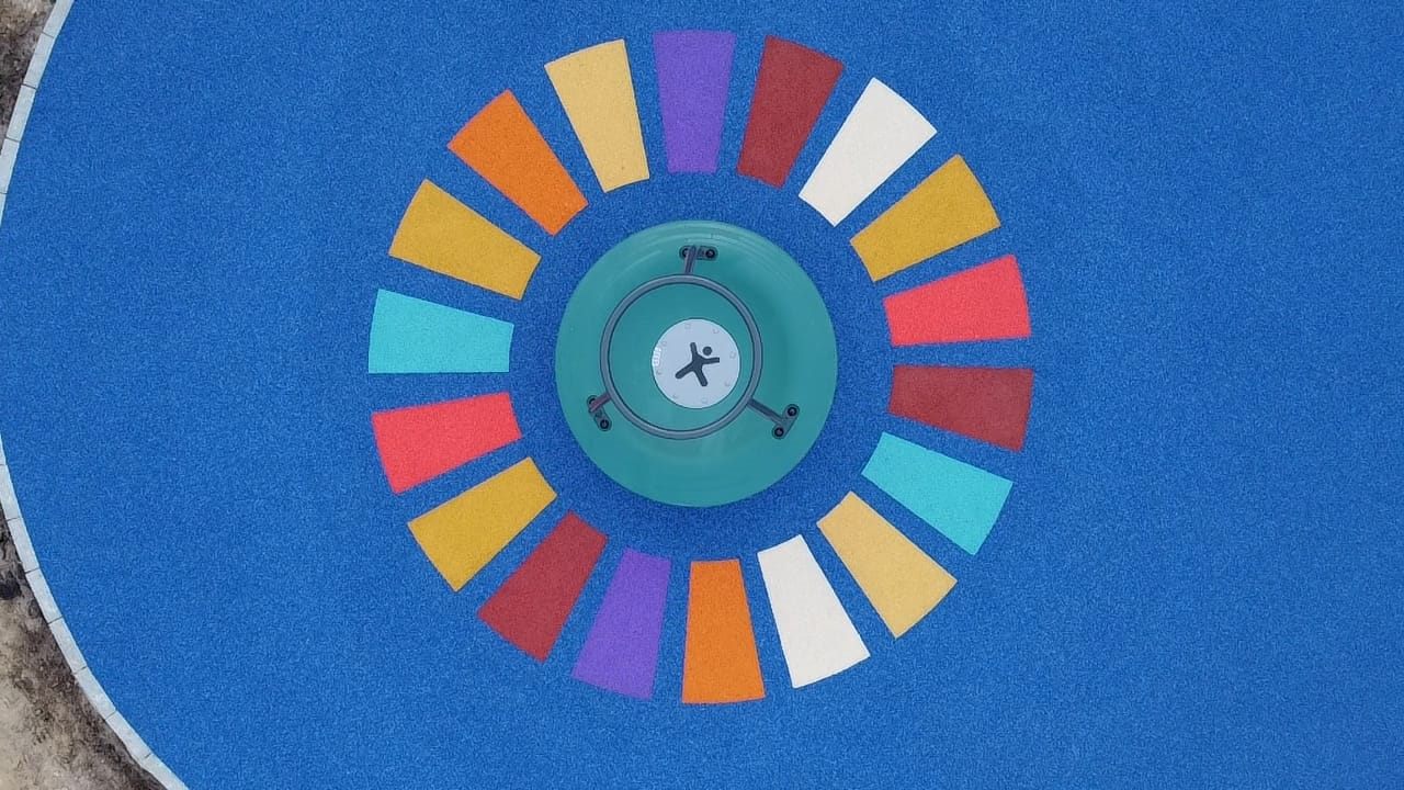 Kreisförmige bunte Intarsie rund um ein Spielgerät auf blauem Fallschutzbelag.