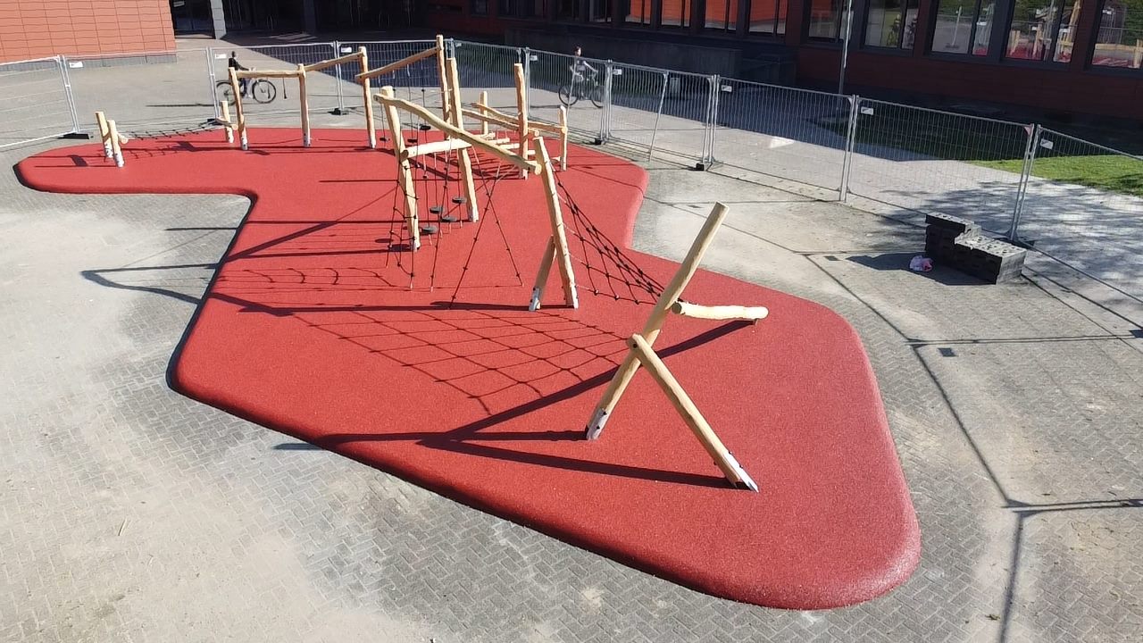 Rote Spielfläche mit Kletter- und Balanciergeräten, umgeben von grauem Pflaster.