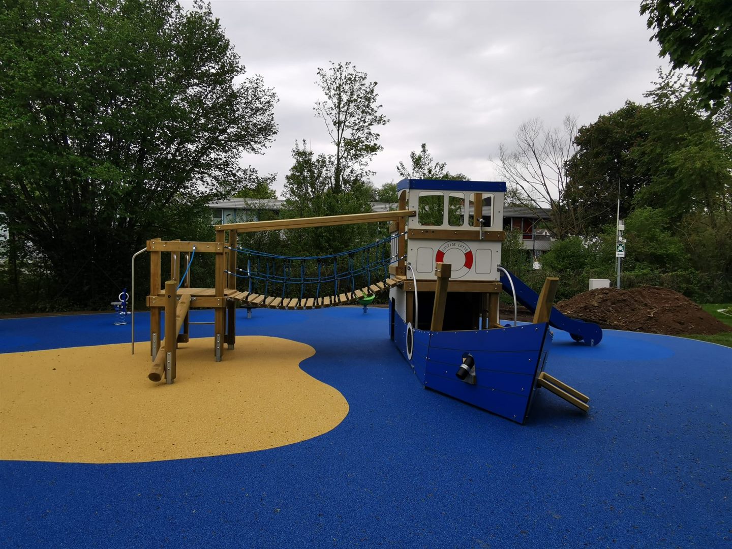 Spielschiff mit gelben und blauen Fallschutzflächen, umgeben von Bäumen.