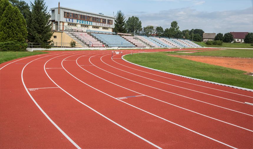 Rote Laufbahn mit weißen Bahnmarkierungen neben einem Sportfeld.